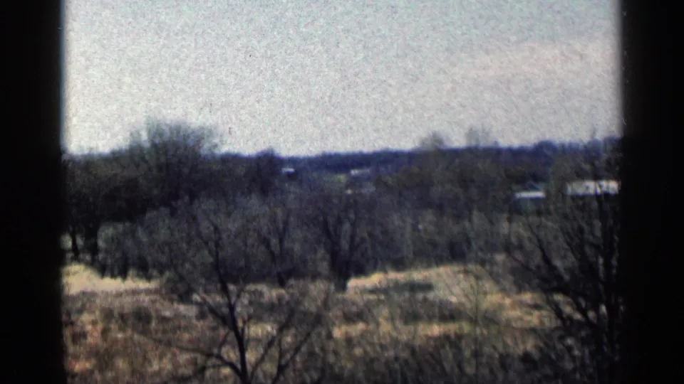 1964 Minnesota 8mm frame showing a rural field, bare trees, small distant buildings, pale sky, and dark vertical edges from the filming position.