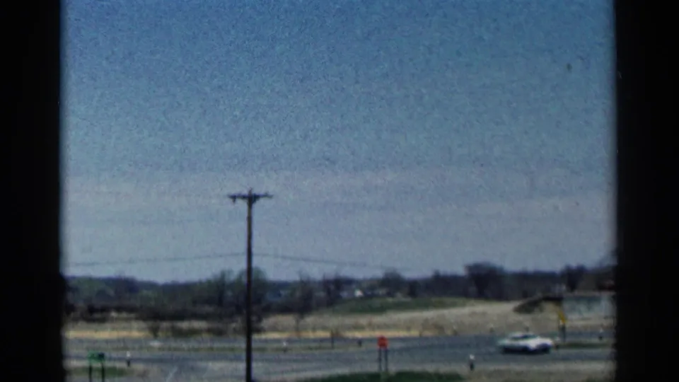 A Minnesota roadside view shows a utility pole, open fields, blue sky, and a small car passing in 1964 home-movie footage.