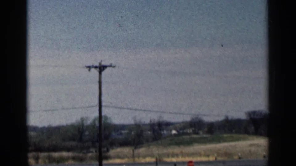 Rural Minnesota roadside in 1964 with a telephone pole, utility wires, fields, bare trees, and small distant buildings.
