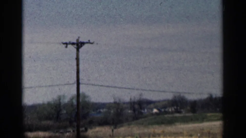 A 1964 Minnesota roadside frame shows a utility pole and wires over open fields, trees, and a low distant horizon.