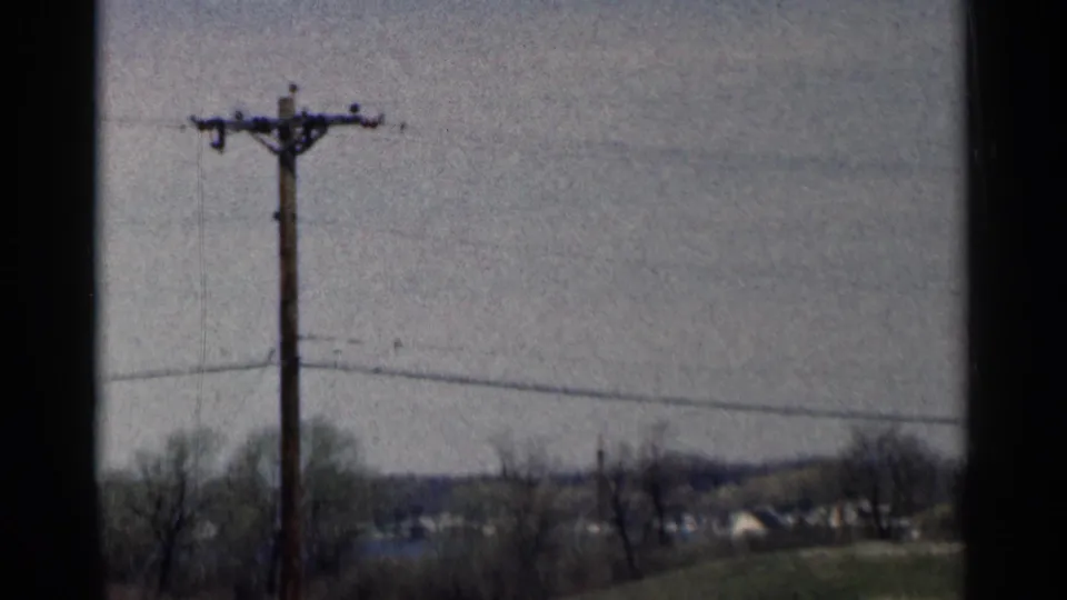 End frame of a 1964 Minnesota rural road view with a close utility pole, crossing wires, fields, trees, and small distant buildings.