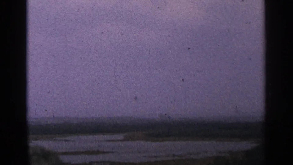 1964 Minnesota 8mm frame with a soft, hazy view over water toward a low shoreline under a cloudy purple-grey sky.