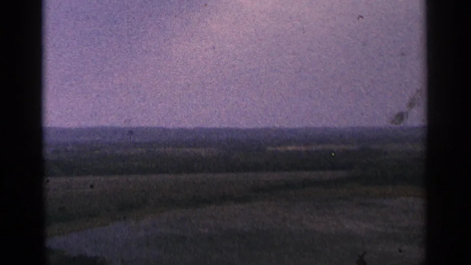 1964 Minnesota 8mm frame showing open fields, a dark horizon line, and distant low hills beneath a muted purple sky.