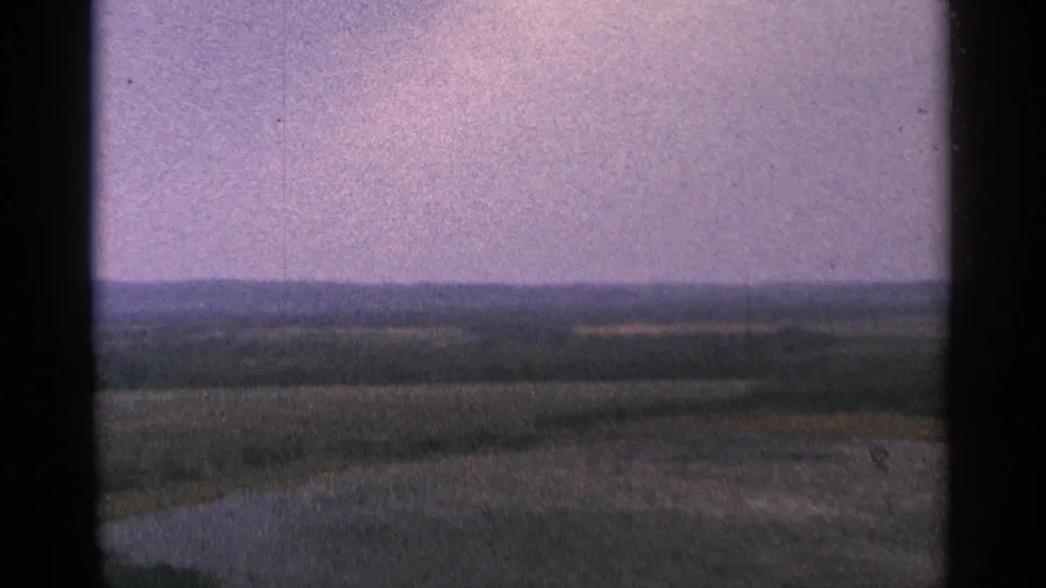 1964 Minnesota 8mm frame with distant hills, layered fields, and a broad hazy sky, partly divided by the scanned film frame.