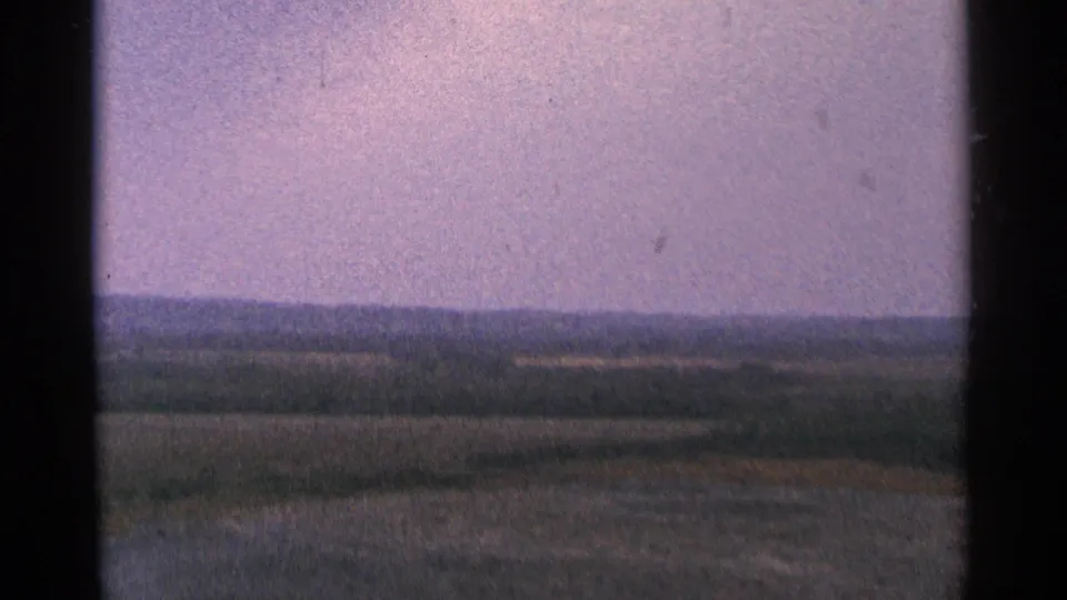 1964 Minnesota 8mm frame showing a wide rural horizon, muted fields, and distant low hills beneath a pale purple sky.