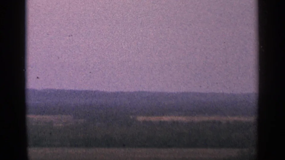1964 Minnesota 8mm frame with the camera zoomed closer toward distant hills, dark field bands, and a muted purple sky.