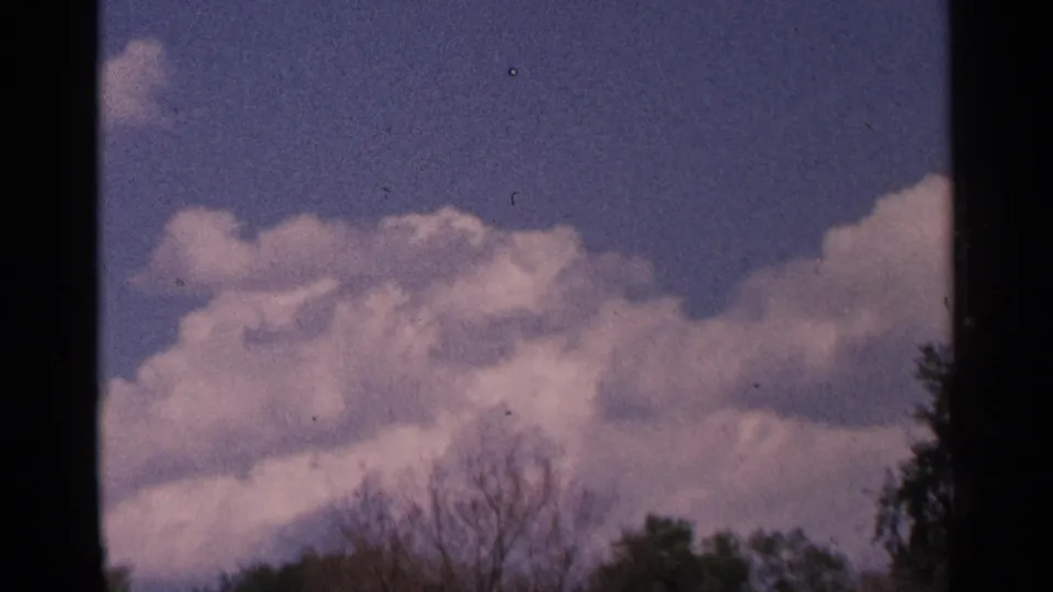 Minnesota 1964 8mm frame looking upward at white clouds in a blue, slightly purple-tinted sky with treetop edges below.