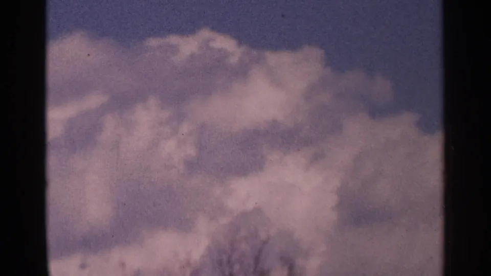 Minnesota 1964 home-movie sky cutaway with layered white clouds, faint branches near the bottom, and visible 8mm film texture.