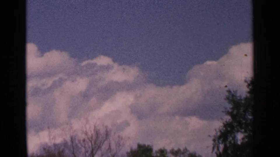 1964 Minnesota 8mm view of cumulus clouds in a blue sky, framed by dark treetop silhouettes and the edge of the film frame.