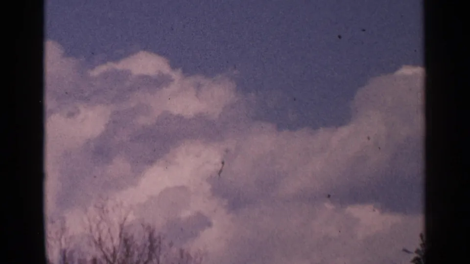 1964 Minnesota 8mm frame centered on bright clouds and blue sky, with bare branches visible near the lower edge.