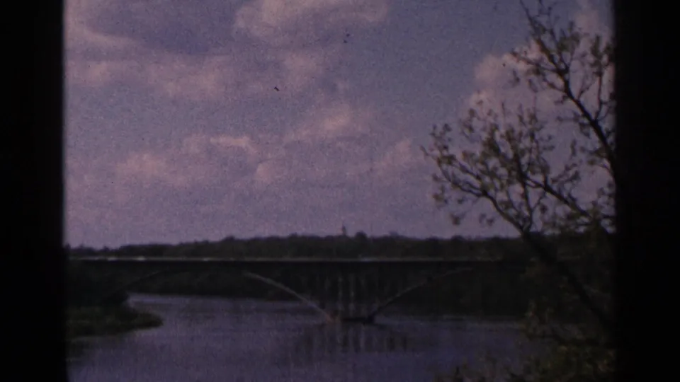 1964 Minnesota 8mm frame showing a distant arched bridge over a river, with sky, clouds, shoreline, and a tree at the right edge.