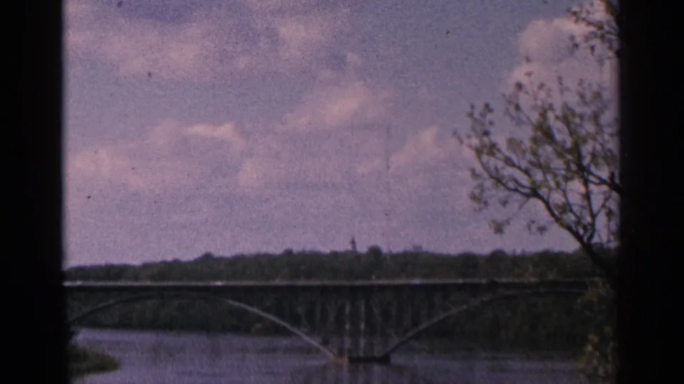1964 Minnesota 8mm view of an arched bridge over a river, framed by dark edges and foreground tree branches.