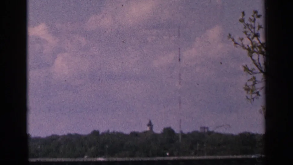 1964 Minnesota 8mm frame showing a radio tower above trees across the river under a cloudy sky.