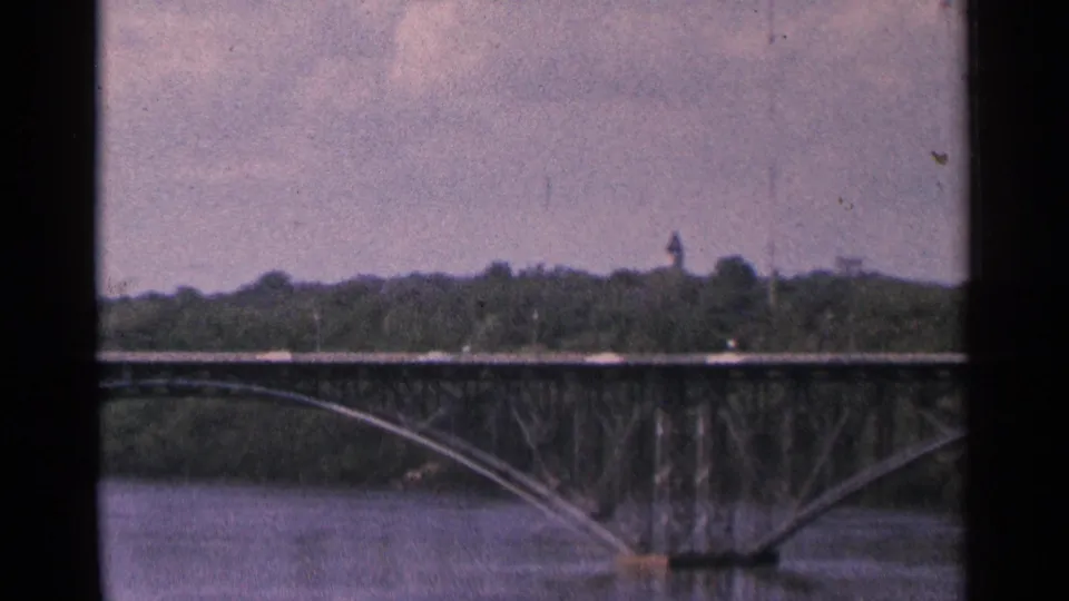 1964 Minnesota 8mm view of a bridge arch over the river with a distant tower visible beyond the treeline.