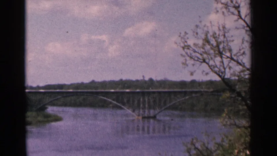 1964 Minnesota 8mm frame with a wide river, arched bridge, distant treeline, and foreground branches at the right edge.