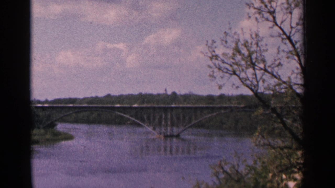 Arched bridge crossing a calm river with trees in the foreground.