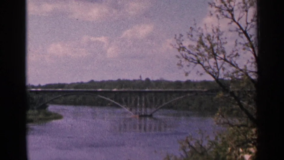 1964 Minnesota 8mm frame showing an arched bridge over still water, partly framed by bare and leafy trees.