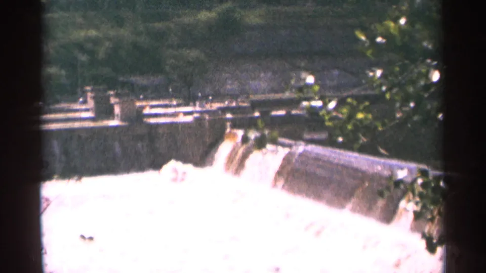 1964 Minnesota 8mm frame showing a low dam or spillway with bright water cascading below a wooded backdrop.