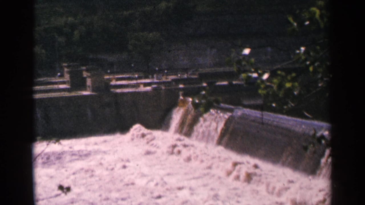 Close view of water rushing over a concrete dam spillway with railings and structures behind it.