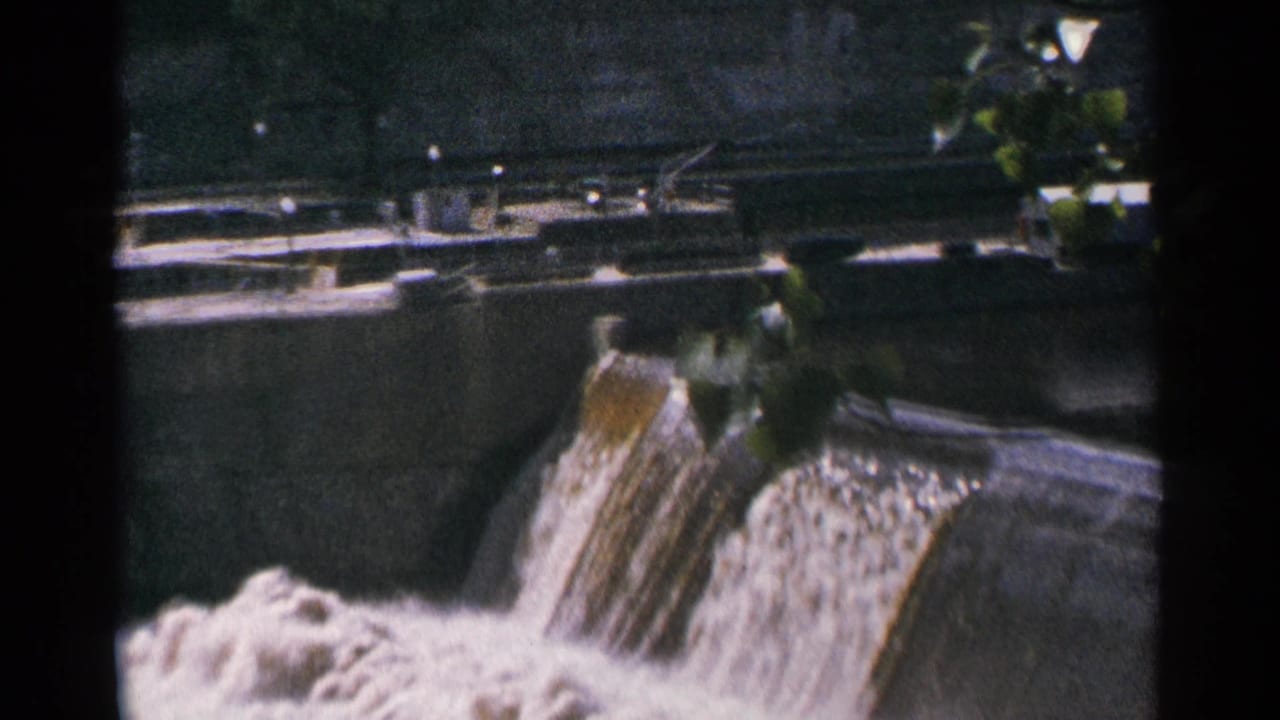 Concrete dam spillway with water falling through a central channel and foliage at the edge of the frame.