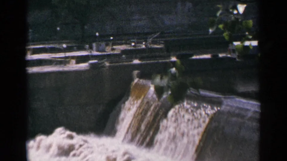 1964 Minnesota home-movie frame of water spilling through a concrete dam channel, partly framed by leaves and backed by railings.