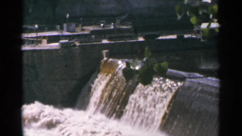 1964 Minnesota 8mm view of a dam spillway from an angle, showing white water below, concrete surfaces, and nearby foliage.
