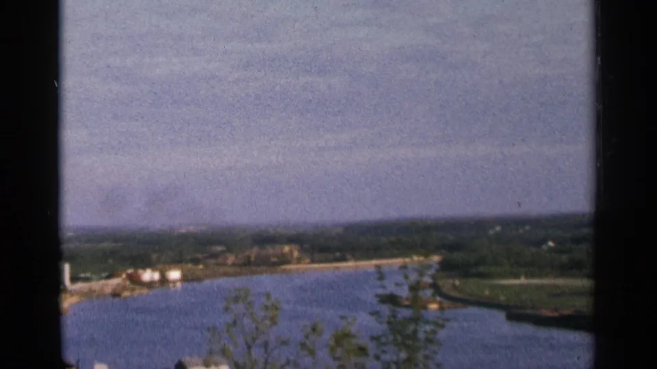 1964 Minnesota 8mm frame widening to a reservoir view, with blue water, distant shore structures, trees, and a hazy horizon.