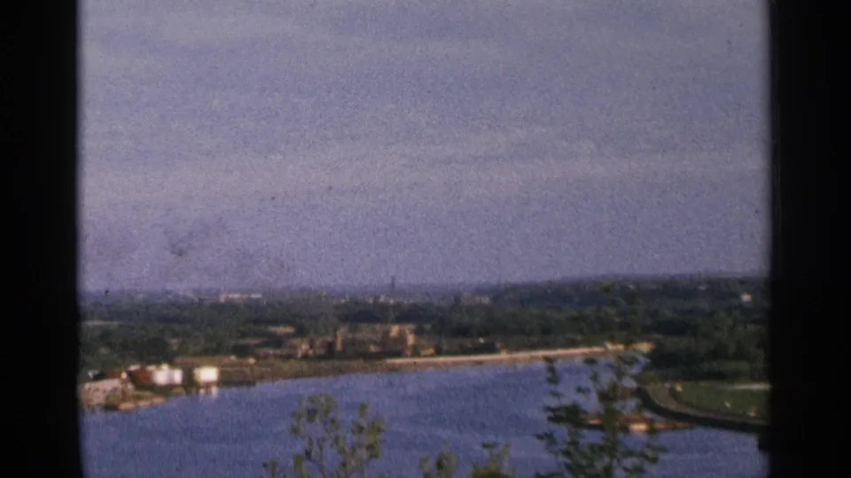 1964 Minnesota 8mm view of a broad river bend with distant city structures, industrial tanks, treetops, and hazy sky.