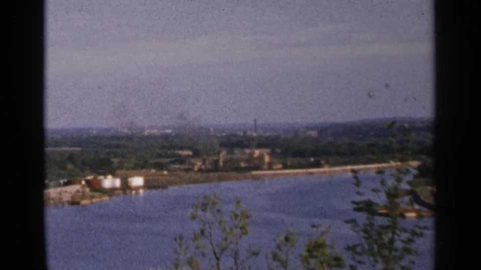 1964 8mm view across a broad Minnesota river bend toward industrial shoreline structures and a faint city skyline.