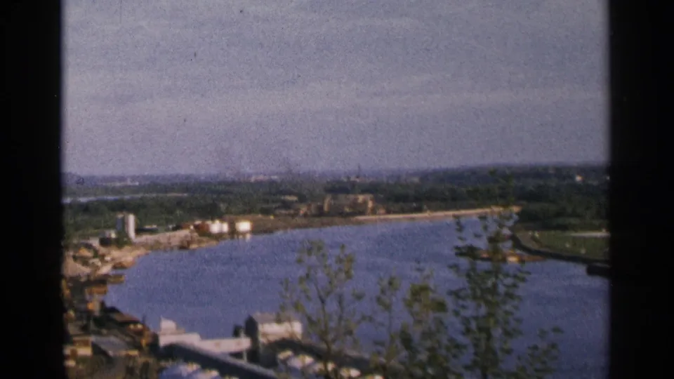 Minnesota home-movie frame showing a blue river curve, treetops, waterfront buildings, and a distant urban background.