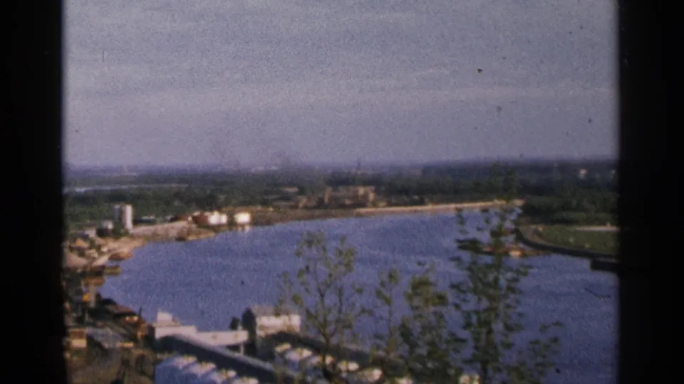 1964 Minnesota 8mm frame showing a broad waterway, wooded shore, and small industrial structures in the distance.