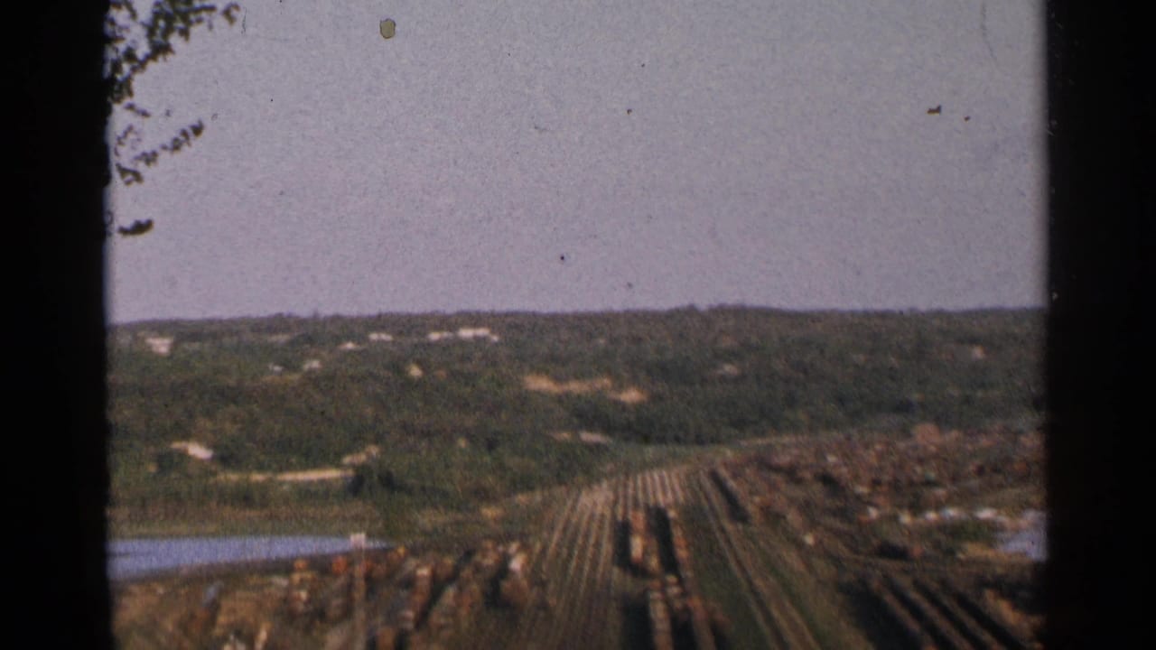 1964 Minnesota 8mm frame showing a lumber yard or log storage area beside wooded terrain and water.