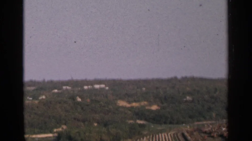 1964 Minnesota 8mm frame looking over a lumber area toward trees, low buildings, and rolling terrain.