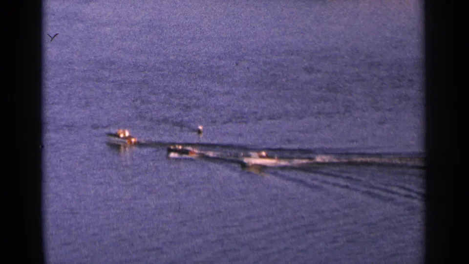 Three small motorboats race across a calm Minnesota lake in 1964 home-movie footage, seen from a distant elevated viewpoint.