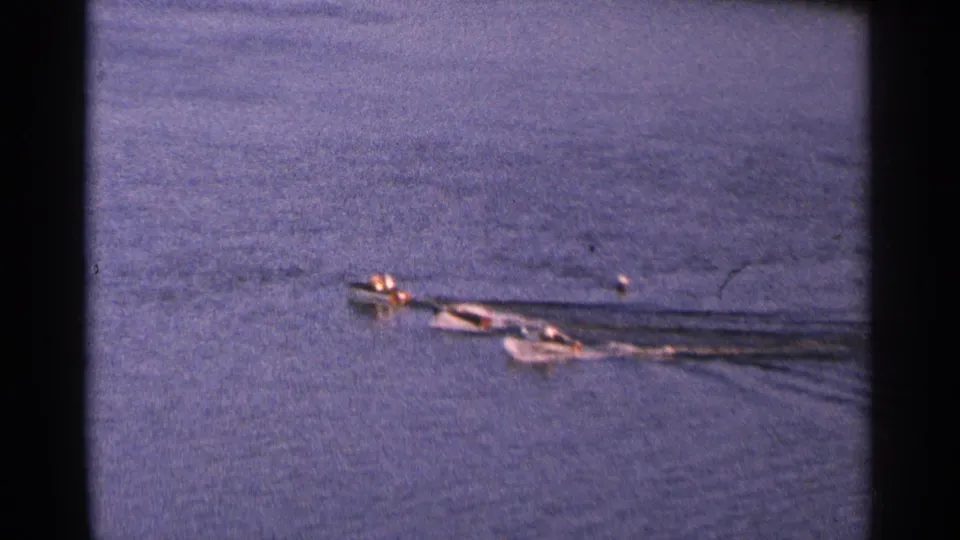 Motorboats continue across the blue lake surface in a 1964 Minnesota 8mm scene, with distant occupants barely visible.
