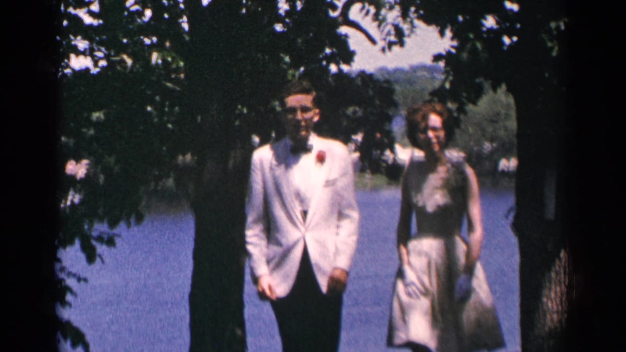 Couple in formal wear walks under trees beside a lake.