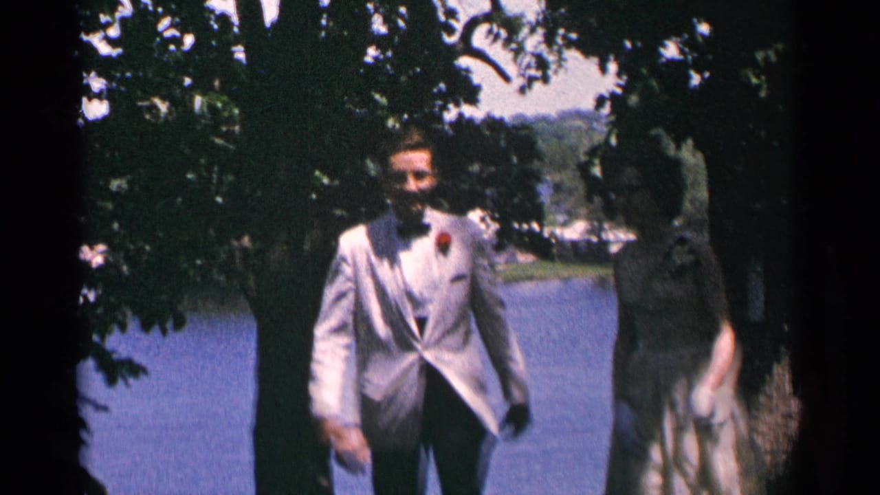 The man in a white formal jacket and boutonniere walks beside a woman in a light dress near a Minnesota lake.