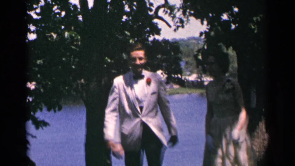 The man in a white formal jacket and boutonniere walks beside a woman in a light dress near a Minnesota lake.