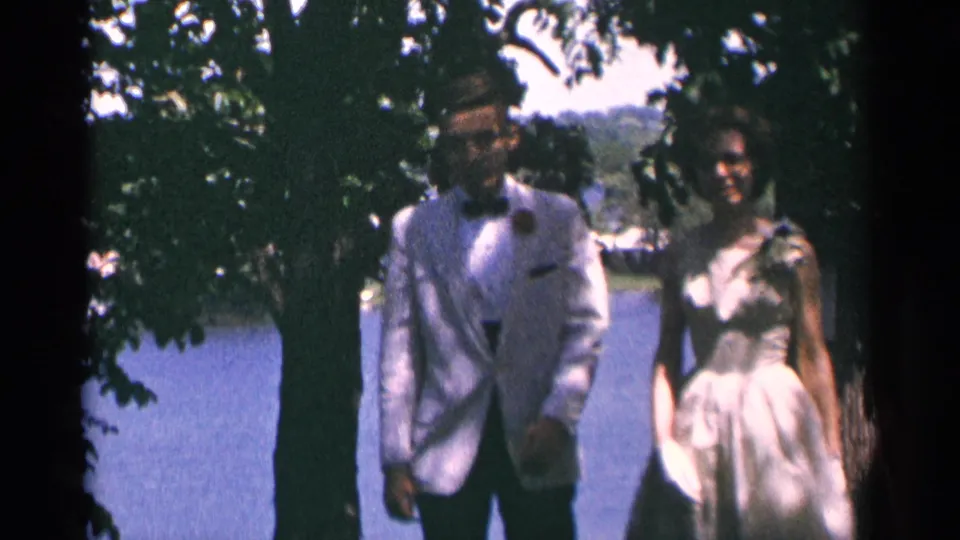A formally dressed couple, likely newlyweds based on the clip metadata, pauses close to the camera beside lake water and trees.