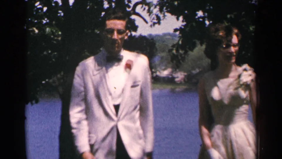 Close lakeside view of the man in a white tuxedo jacket and the woman in a light dress walking through shade in 1964 Minnesota footage.