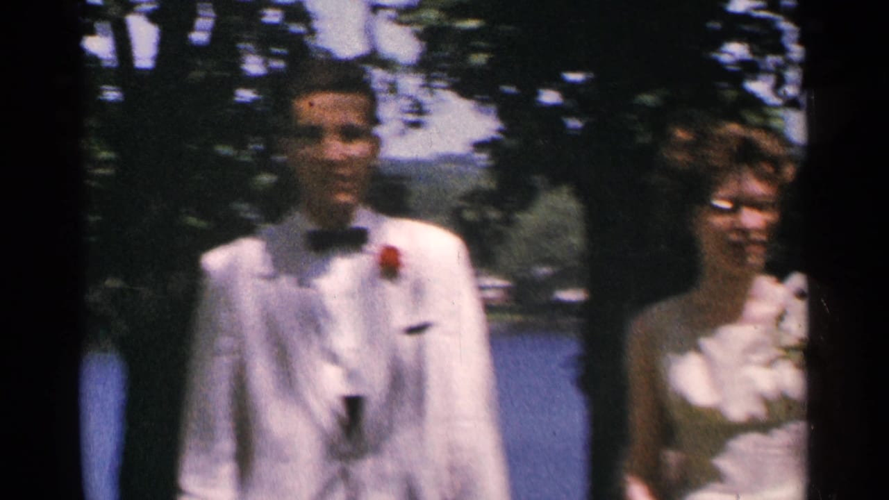 Young couple in formal attire posing outdoors beside trees and water.