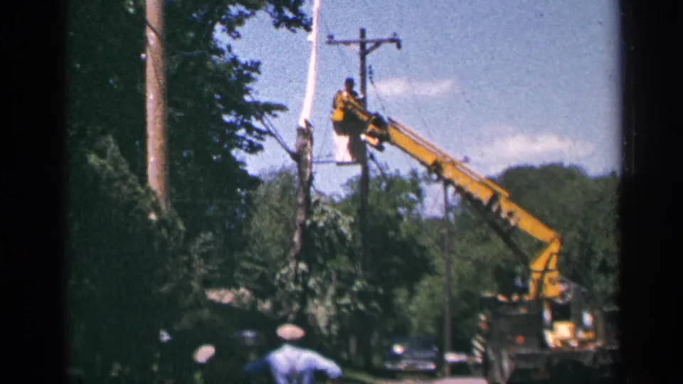 The raised crane bucket holds a utility worker near wires while people and older cars appear on a tree-lined Minnesota street.