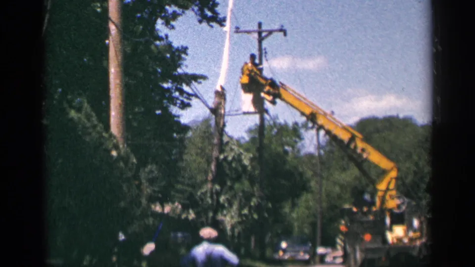 A yellow crane and bucket remain positioned at a utility pole on a leafy Minnesota street, with parked cars and onlookers below.