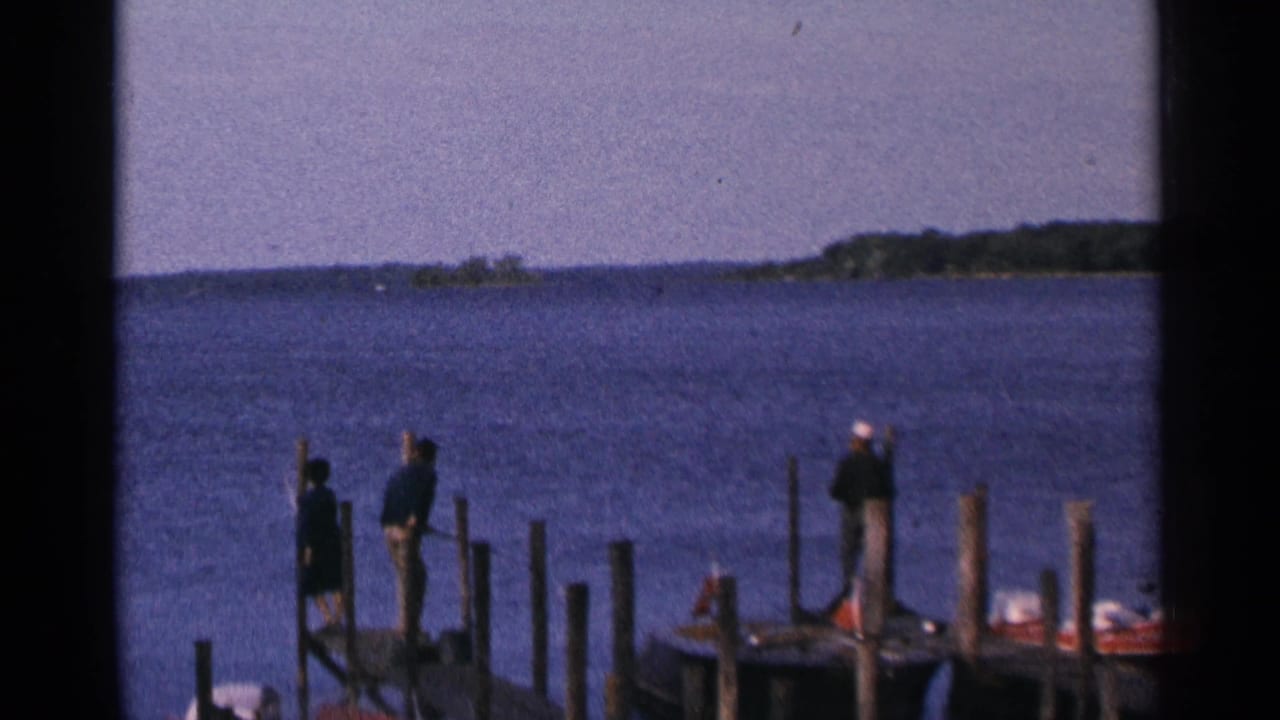 People stand on wooden piers beside a broad lake with a distant tree-lined shore.