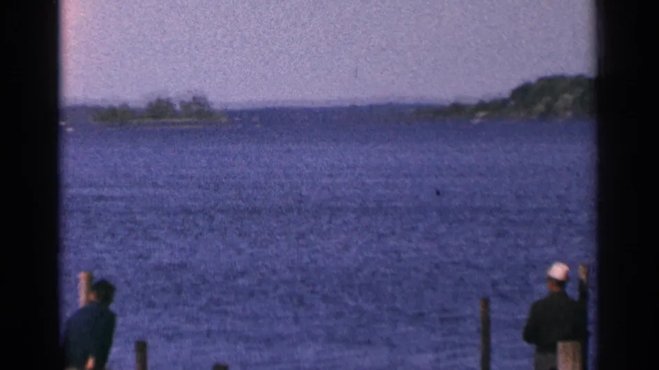 1964 Minnesota lakefront view with a person near dock pilings and open water stretching toward tree-covered land.