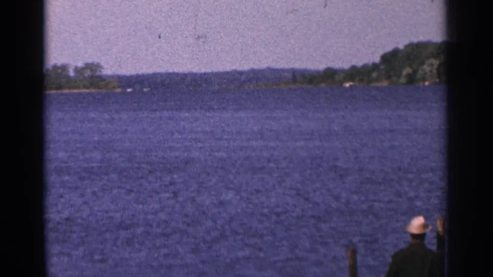 1964 Minnesota home-movie view across a large lake, with wooded shoreline and a partial person visible near the dock edge.