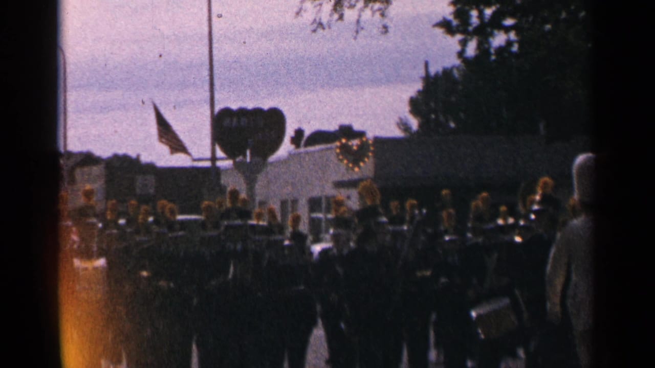 Marching band in dark uniforms passes a decorated float and flag.