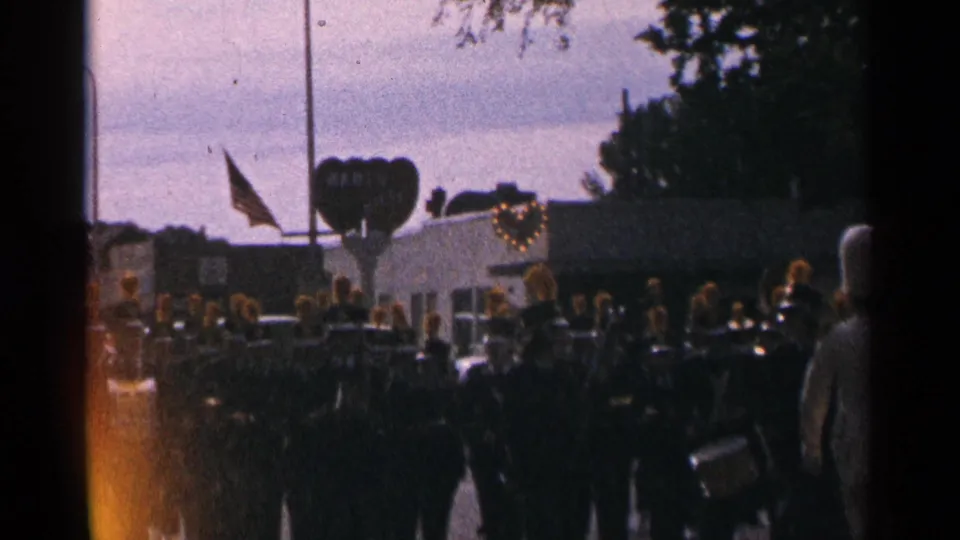 1964 Minnesota parade view of a marching band in dark uniforms with gold plumes near a flag and heart-shaped float.
