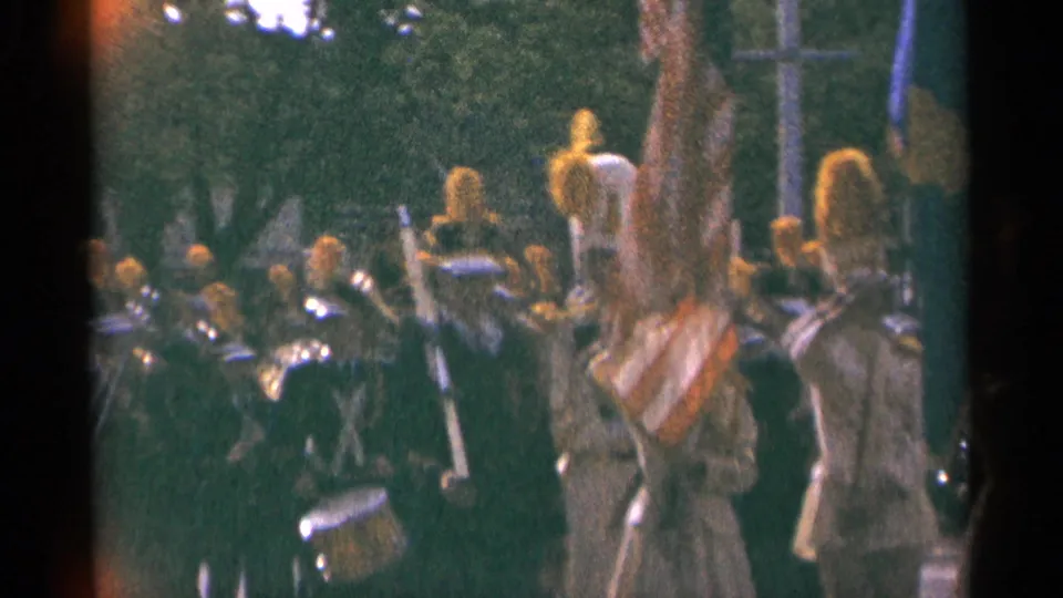 Handheld 8mm frame of a Minnesota parade band with drummers, brass players, gold plumes, and a flag crossing the foreground.