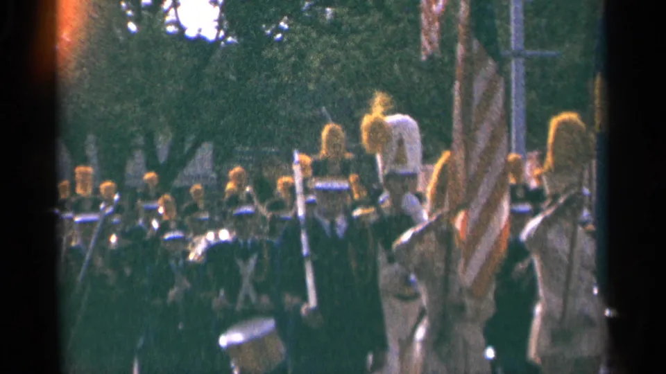 Minnesota parade footage shows uniformed band members carrying drums and brass instruments beneath roadside trees.